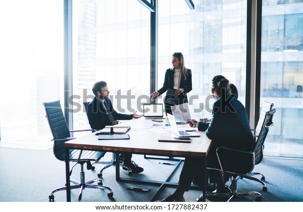 High angle of content female entrepreneur\
in elegant clothes explaining business strategy to board of\
directors while standing near table during\
meeting