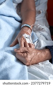 High angle of crop anonymous elderly man holding hand of sick woman with connected venous catheter lying on bed during rehabilitation treatment in hospital