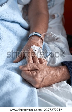 High angle of crop anonymous elderly man holding hand of sick woman with connected venous catheter lying on bed during rehabilitation treatment in hospital