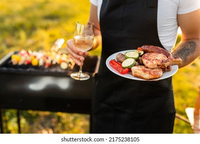 High angle of crop unrecognizable waiter in black apron serving glass of white wine and plate, with grilled veggies and meat during event in park on sunny day - Powered by Shutterstock