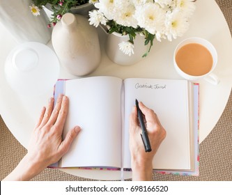 High angle cropped view of woman's hands writing in gratitude journal at desk with tea and flowers (selective focus)