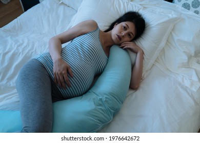 High Angle Depressed Pregnant Mom Lying Alone Awake With A Maternity Pillow On Bed Is Looking Into Space While Suffering Insomnia In The Bedroom At Midnight.