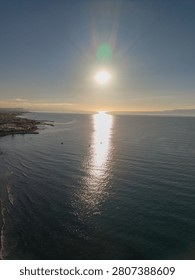 High angle drone perspective captures a bright sun setting over the sea surface creating a strong golden light reflection near a coastal shoreline.