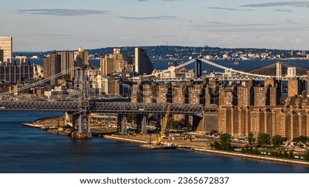 A high angle drone shot looking south over the East River in New York on a gray and cloudy day. The Williamsburg, Manhattan and the Brooklyn Bridges are visible in the background distance.