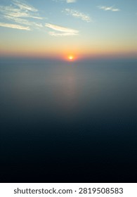 High angle drone view of a glowing sun at sunrise, or sunset over the endless open ocean horizon with warm golden light reflecting on calm ripples.