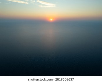 High angle drone view of a glowing sun at sunrise, or sunset over the endless open ocean horizon with warm golden light reflecting on calm ripples.