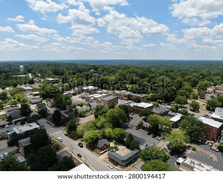 High angle drone view of the historic downtown district in Wake Forest, North Carolina, featuring brick retail storefronts, streets with parking, municipal buildings, and lush green trees under a bright blue summer sky with light clouds.