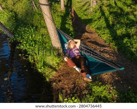 High angle drone view of peaceful blonde woman sleeping in green hammock hung on shore of calm forest lake during summer vacation