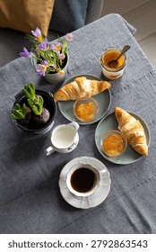High angle flat lay of a dark linen table showing a cup of black coffee, glass jar of orange jam, fresh croissants on plates, and pots of purple crocus flowers.