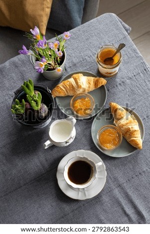High angle flat lay of a dark linen table showing a cup of black coffee, glass jar of orange jam, fresh croissants on plates, and pots of purple crocus flowers.