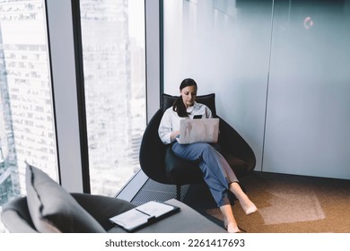 High angle full body of concentrated female manager in elegant outfit sitting in armchair and working on business project while browsing laptop at modern workplace