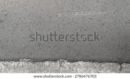 High angle full frame texture view of a freshly paved dark gray asphalt road surface bordering an old weathered concrete edge block. The horizontal division showcases a distinct contrast between coarse textures, providing a clean rustic background template for urban infrastructure layout presentation concepts.