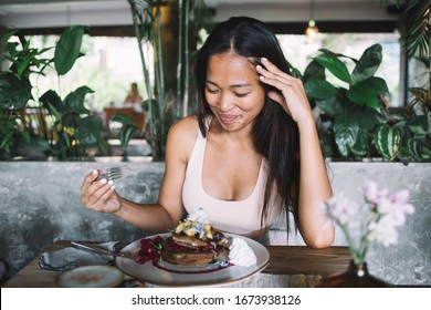 High Angle Of Gorgeous Asian Embarrassed Female Looking Down And Laughing While Sitting At Table And Eating Tasty Dessert In Modern Cafe