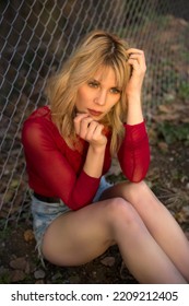 High Angle Of Gorgeous Young Female Model In Mini Denim Shorts And Red Blouse Touching Long Blond Hair And Looking Away Near Chain Link Fence In Countryside