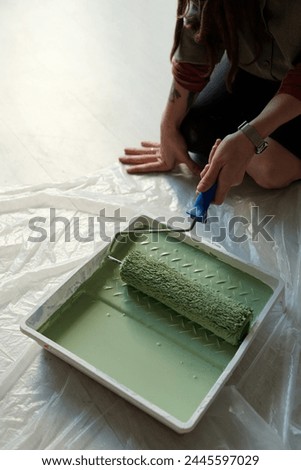 High angle of hand of young female worker sitting on knees on the floor covered by cellophane and putting paintroller in tray with green paint