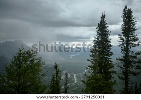 High angle landscape view of Bow Valley and distant mountain ranges under dark storm clouds with rain curtain, framed by tall pine trees in Banff National Park, Alberta, Canada.