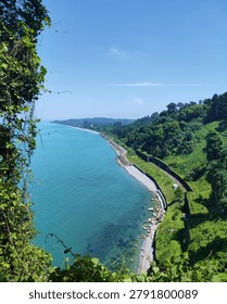 High angle look down view of a scenic coastline with a railway line passing through green tropical hills next to a clear turquoise sea