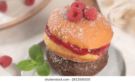A high angle macro shot captures two sugar coated donuts stacked on a plate one filled with red jam and topped with fresh raspberries Bright soft studio lighting enhances the rich sweet textures