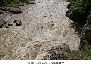 High angle medium shot capturing the pure chaos, roaring sound, and extreme danger of a raging mountain torrent. The muddy brown water of the Belaya River in Adygea, Caucasus, churns violently, forming dangerous standing waves, hydraulic drops, and crushing foam whirlpools. This scene illustrates the devastating kinetic energy, flash floods, and massive sediment load causing rapid channel erosion. Perfect for commercial media depicting environmental hazards, natural disaster reports, survival guides, and high-risk extreme adventure sports