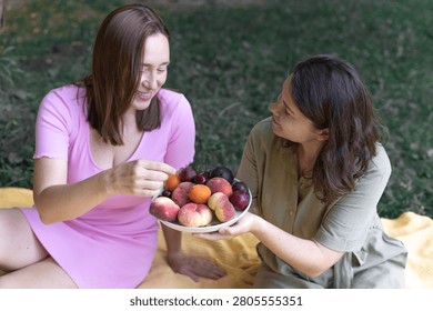 High angle medium shot of two women sitting on a blanket in the grass, holding and sharing a plate of fresh peaches, plums, and cherries. Bright, sharp, detailed.
