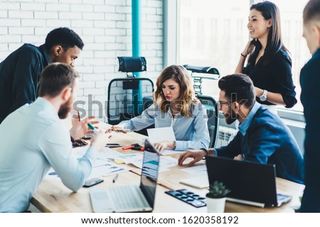 High angle of multiracial business partners in elegant clothes listening to coworker proposition while examining reports and using digital devices at desk in light contemporary conference room Foto stock © 