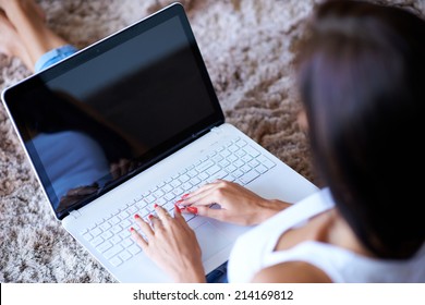 High Angle Over The Shoulder View Of The Hands Of A Woman Typing On A Laptop Computer As She Sits On The Carpet At Home