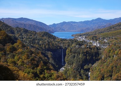 High angle panoramic landscape view of the world-famous Kegon Falls (Kegon no Taki) cascading from a towering cliff, with the serene Lake Chuzenji and the lush forest of Nikko National Park in the background during the peak autumn season. This spectacular vista captured from Akechidaira Plateau showcases the vibrant red, orange, and yellow autumn foliage (koyo) against the deep blue waters of the lake and the distant mountain ranges under a clear sunny sky. A quintessential autumn scenery of Tochigi Prefecture, Japan, highlighting the natural beauty and geological wonders of the Kanto region, perfect for travel, tourism, and nature-themed commercial projects.