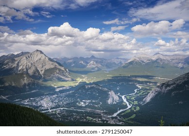 High angle panoramic landscape view of Banff town, the winding Bow River, and the majestic Canadian Rockies mountain range under a blue cloudy sky in Alberta, Canada.