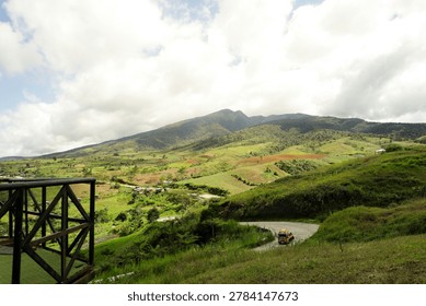 High angle panoramic view of a winding mountain road with a driving truck, lush green rolling hills, agricultural fields, and a majestic mountain range under a cloudy sky.