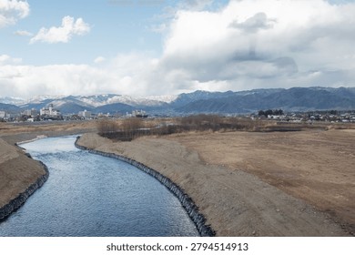 High angle panoramic view of a winding blue river flowing through a rural valley with a majestic snow capped mountain range under cloudy skies in Japan
