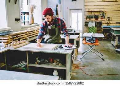 High angle of pensive diligent carpenter in plaid shirt and gray apron concentrating and drawing with pencil on plank while standing at table with blueprint and tools in comfortable modern workroom - Powered by Shutterstock