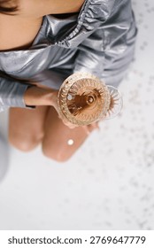 High angle perspective of a lady sitting on a white floor, holding a crystal coupe glass with sparkling wine. Scattered confetti on the floor adds to the festive holiday mood.