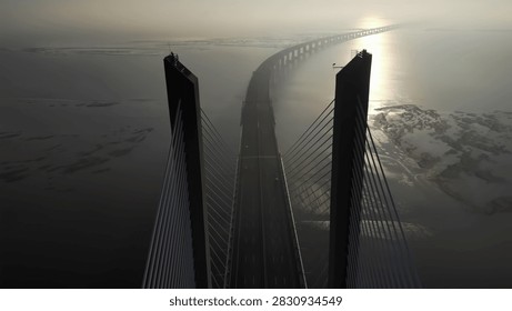 High angle perspective of long cable stayed bridge crossing over water at sunrise. Golden light reflects on surface with misty atmosphere and abstract architectural lines.
