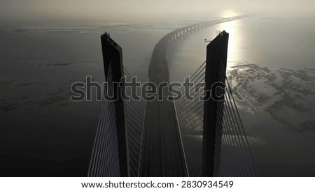 High angle perspective of long cable stayed bridge crossing over water at sunrise. Golden light reflects on surface with misty atmosphere and abstract architectural lines.