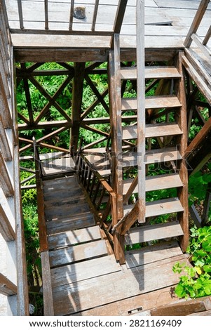 High angle perspective view of a wooden lookout tower staircase with intricate cross bracing and lush green forest foliage background.