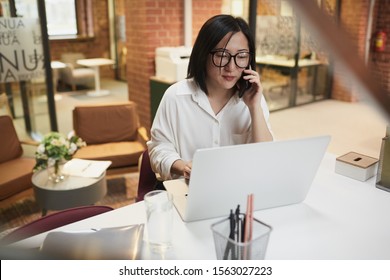 High angle portrait of successful Asian businesswoman sitting at workplace in modern office and speaking by phone while managing deal, copy space