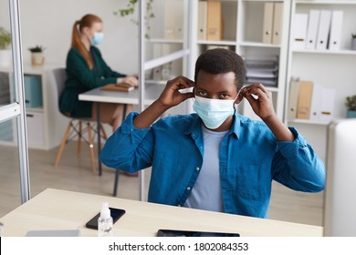 High Angle Portrait Of Young African-American Man Putting On Face Mask And Looking At Camera While Working At Desk In Post Pandemic Office