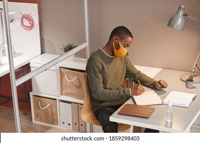 High Angle Portrait Of Young African-American Man Wearing Mask And Writing In Planner While Working At Desk In Office Cubicle, Copy Space