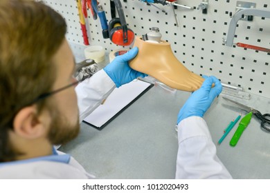 High Angle Portrait Of Young Doctor Holding Artificial Foot At Desk In Office, Checking It For Quality And Making Adjustments