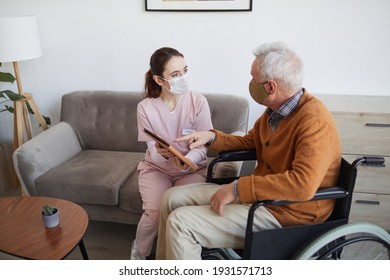 High angle portrait of young female nurse assisting senior man in wheelchair using digital tablet at retirement home, both wearing masks, copy space - Powered by Shutterstock