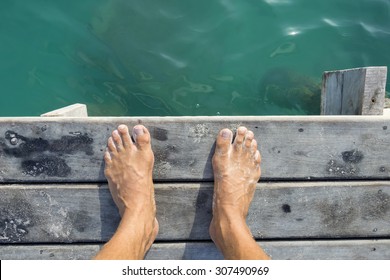 High Angle POV Of Man's Bare Feet Standing At Edge Of Wooden Dock Sprinkled With Fine White Sand Above Aqua Green Sea Water In Natural Morning Light