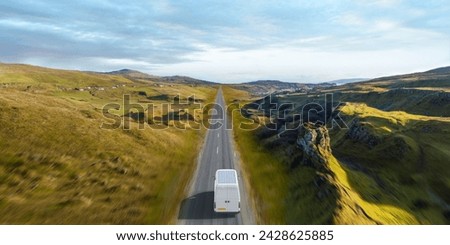 A high angle, rear view of a generic electric white van with solar panels fitted to the roof driving through green, hilly countryside on an open road in bright weather. Motion blur to foreground