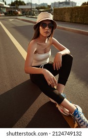 High angle of self assured young female millennial in casual outfit, with panama hat and sunglasses sitting on asphalt road on sunny day in city