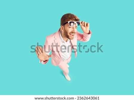 High angle shot from above of happy cheeky handsome young man wearing pink suit standing on blue color background looking at camera, smiling, taking off sunglasses and winking his eye. Fashion concept