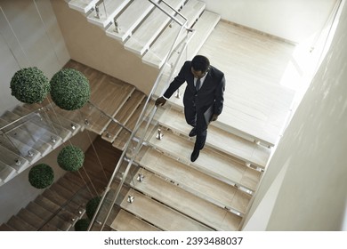 High angle shot of African American businessman wearing suit carrying laptop descending stairs holding on banister in modern office block, copy space - Powered by Shutterstock