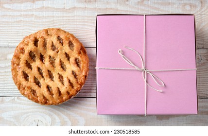 High Angle Shot Of An Apple Pie And A Pink Bakery Box. Horizontal Format On A Rustic White Wood Kitchen Table. 