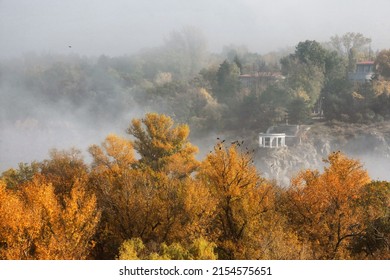 A High Angle Shot Of Autumn Treetops Against A Hill Covered With Greenery And Houses In The Fog