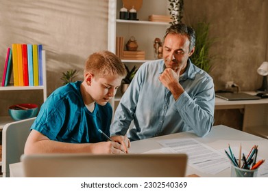 High angle shot of a blond teenage boy and his middle aged father sitting at the dinning table at home while the boy finishing his homework. Supportive father watching his work with joy. Copy space. - Powered by Shutterstock