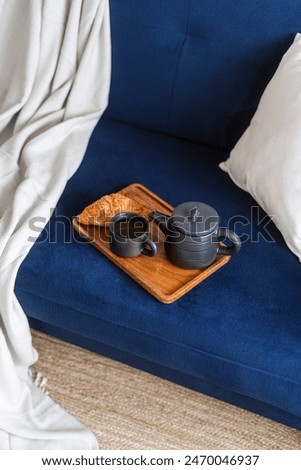High angle shot of ceramic teapot with preparing drink, cup of hot tea and fresh french croissant on breakfast wooden tray on blue comfortable couch with plaid and decorative pillow in living room
