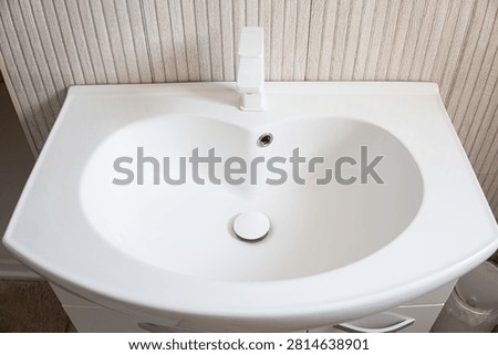 High angle shot of a clean white porcelain bathroom washbasin featuring a sleek faucet, central drain plug, and overflow hole set against light neutral wall tiles.